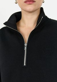 Svart zip-up sweatshirt med hög krage, silverdragkedja och präglad logodetalj på bröstet. Slät textur, avslappnad design.