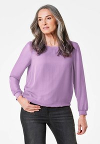 Lila Langarmbluse mit Rundhalsausschnitt, glatter Textur und gerafften Manschetten, kombiniert mit dunklen Jeans. Die Passform ist lässig und entspannt.