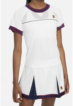 Kvinde iført hvidt tennisoutfit med marineblå og røde kanter, der består af en kortærmet top og nederdel med plisser og meshindsatser.