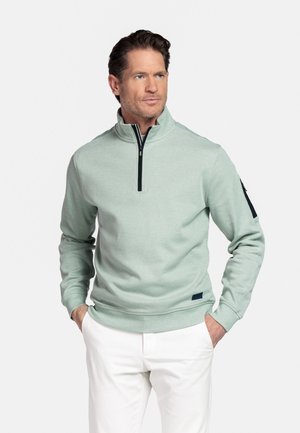 Man draagt een lichtgroene trui met een kwart rits en witte broek, staat met handen in de zakken en kijkt naar links tegen een effen achtergrond.