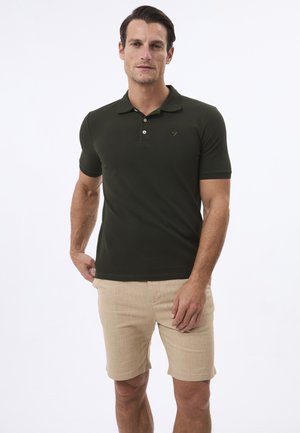Mand iført en mørkegrøn poloskjorte med korte ærmer og beige shorts, der står foran en ensfarvet hvid baggrund.