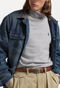 Chaqueta vaquera con cuello de pana, suéter gris de cuello alto y pantalones plisados beige, complementados con un cinturón de cuero marrón.