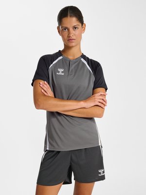 Młoda kobieta ze skrzyżowanymi ramionami ubrana w czarno-szary strój sportowy Hummel, stojąca na jednolitym białym tle.