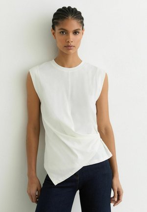 Fonott hajú nő ujjatlan fehér felsőt, aszimmetrikus szegéllyel, és sötét farmert visel, fehér falnak támaszkodva.