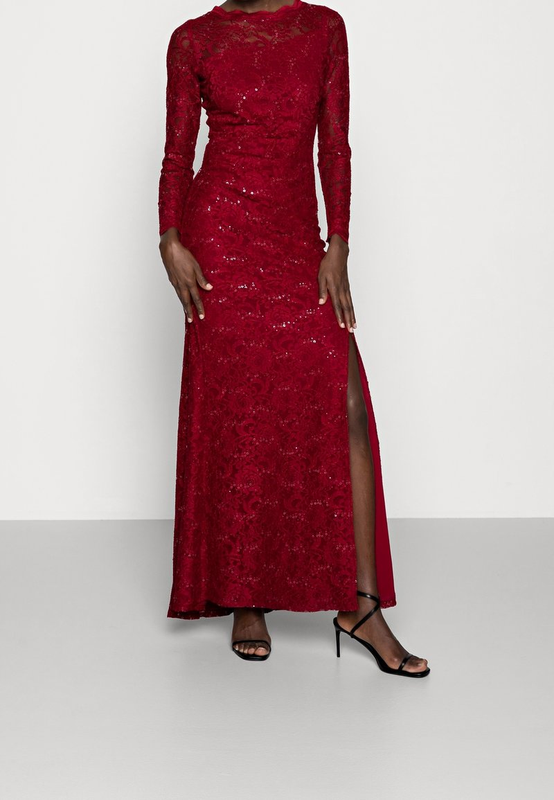 Femme portant une robe en dentelle rouge à manches longues avec des sequins et une fente haute, associée à des sandales noires à talons hauts à brides.