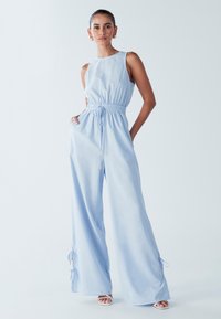 Svetlo modra kombineža brez rokavov, z nabranim pasom in širokimi hlačnicami. Nastavljive vezalke na gležnjih; mehka, lahka tkanina.