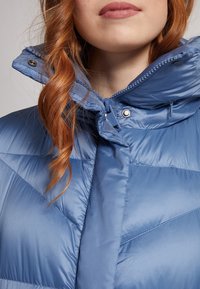 Gros plan sur une personne aux cheveux roux portant une veste matelassée bleue avec un col montant zippé et des boutons pressions.