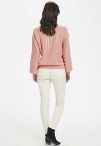 Frau mit welligem braunem Haar, die einen rosa gestrickten Pullover mit Pailletten, eine cremefarbene Hose und schwarze Stiefeletten trägt, steht vor einem weißen Hintergrund.