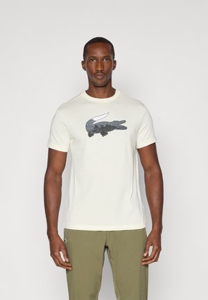 Camiseta de color crema con un gráfico de cocodrilo negro en la parte frontal. Mangas cortas y cuello redondo, combinada con pantalones verde oliva.