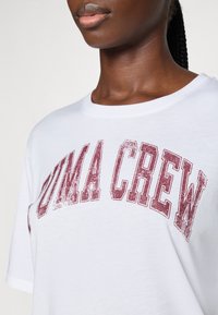 Camiseta de algodón blanca con un estampado gráfico rojo "PUMA CREW" en el pecho, con cuello redondo y mangas cortas.