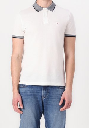 Mand iført hvid polo med korte ærmer, grå krave og manchetter, lille logo på brystet, og blå jeans, står på en hvid baggrund.