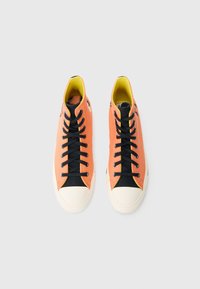 Υψηλά αθλητικά παπούτσια με πορτοκαλί επάνω μέρος από καμβά, μαύρες λεπτομέρειες, μαύρα κορδόνια και λευκή λαστιχένια μύτη. Κίτρινη εσωτερική επένδυση, λογότυπο στην πλευρά.