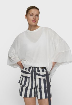 Mujer con el cabello recogido que lleva una blusa blanca holgada y pantalones cortos de rayas verticales negras y blancas de talle alto con grandes bolsillos.