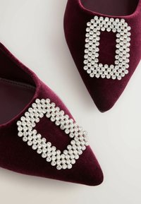 Pāris bordo samta kurpes ar smailiem purngaliem un lielām taisnstūrveida strass sprādzēm uz gaišas virsmas.