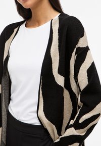 Γυναίκα ντυμένη με μια μαύρη και μπεζ κάρντιγκαν με αφηρημένο σχέδιο, φορεμένη πάνω από ένα λευκό πουκάμισο και μαύρο παντελόνι, απεικόνιση από το λαιμό έως τους γοφούς σε λευκό φόντο.