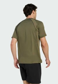 Camiseta atlética de manga corta en verde oliva con detalles negros, textura suave, corte entallado y un sutil detalle de logo en la parte posterior. Se ven pantalones cortos negros.