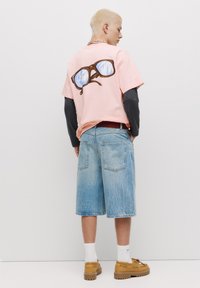Tricou roz supradimensionat cu un grafic cu ochelari de soare supradimensionați pe spate, asortat cu șorturi din denim ușor bleumarin și încălțări bej. Socote albe vizibile.