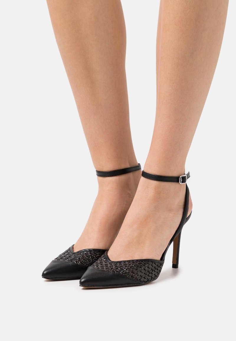 Jambes portant des escarpins noirs à bout pointu avec bride à la cheville et détail supérieur tissé texturé sur un fond clair.
