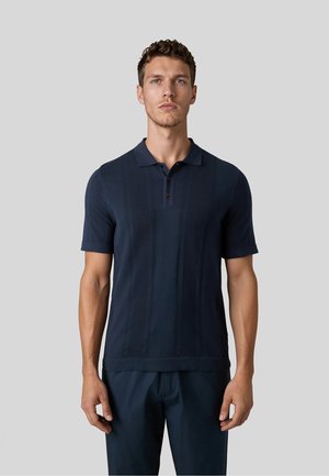 Junger Mann, stehend und nach vorne blickend, trägt ein dunkelblaues Polo-Shirt mit kurzen Ärmeln und passende dunkle Hose vor einem einfarbigen grauen Hintergrund.