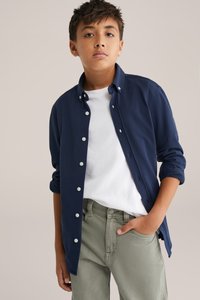 Jūras spēku pogātā krekls ar baltām pogām, uzvilkts virs baltā T-krekla un apvienots ar gaiši zaļām biksēm. Gluda auduma, klasiskā piegriezuma.