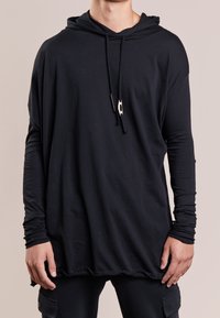 Musta huppari, jossa pitkät hihat ja sileä kangas. Hupussa nyörikiristys ja helmassa kaareva muoto, jossa kerrostettua tekstuuria alhaalla.