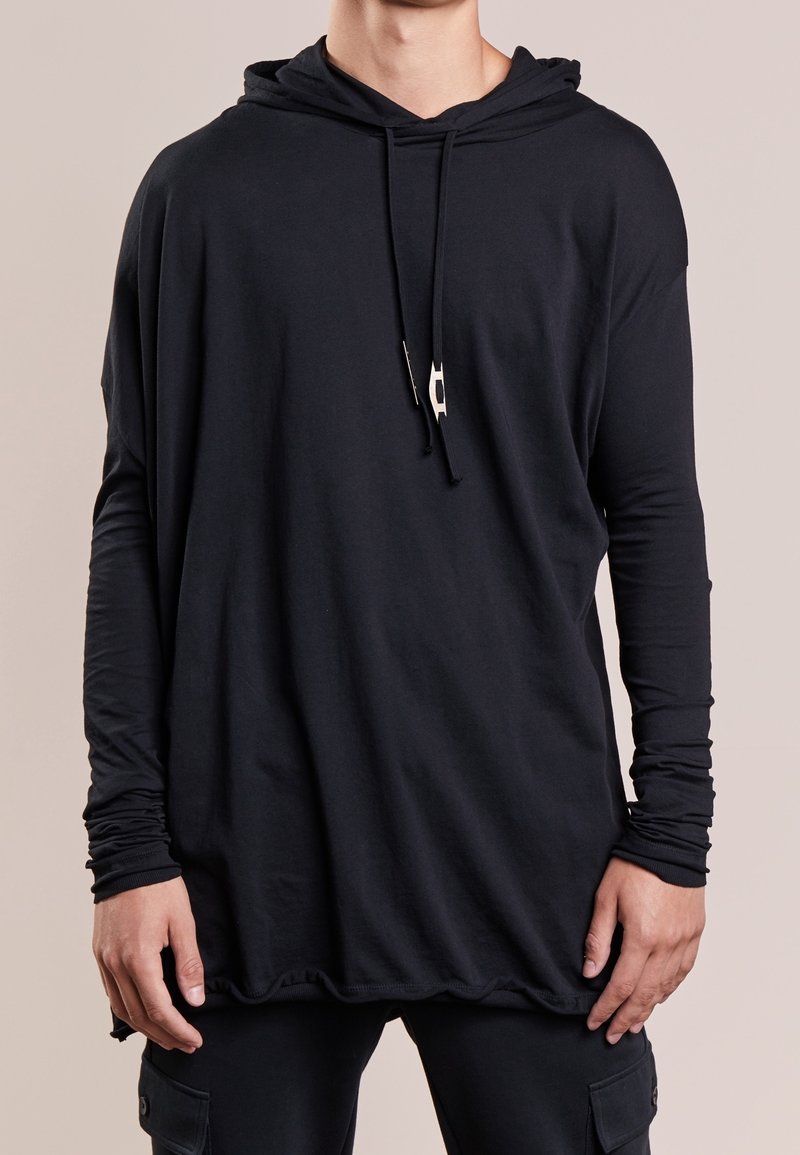 Musta huppari, jossa pitkät hihat ja sileä kangas. Hupussa nyörikiristys ja helmassa kaareva muoto, jossa kerrostettua tekstuuria alhaalla.