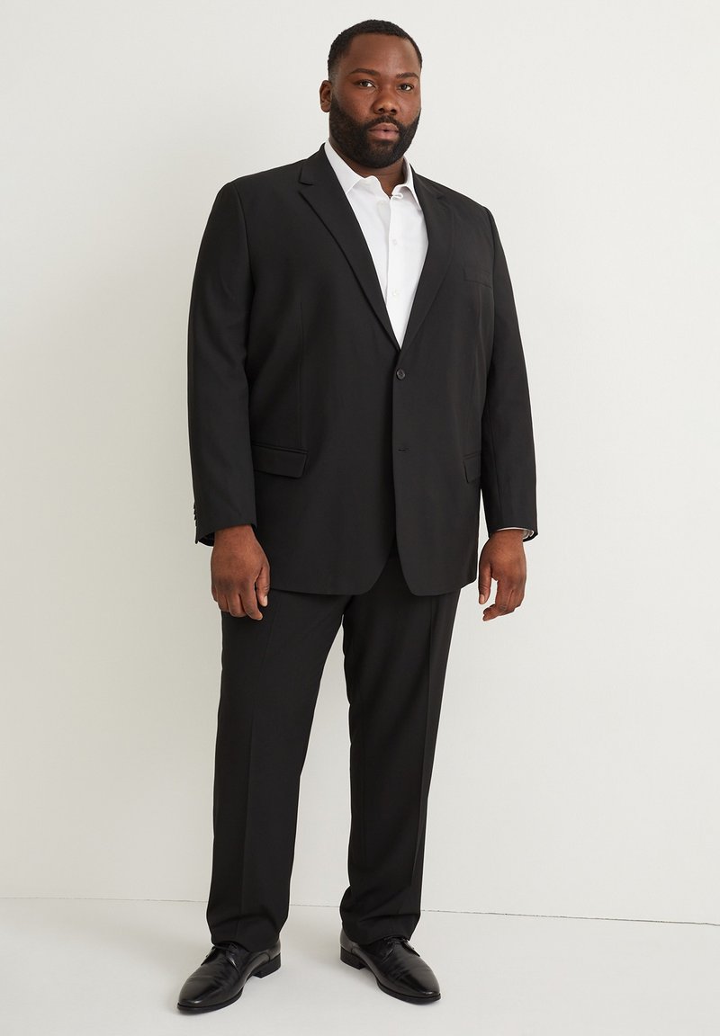 Traje negro con chaqueta de un solo botón y pantalones a juego. Camisa blanca de vestir debajo. Tejido suave y corte moderno. Zapatos negros brillantes.