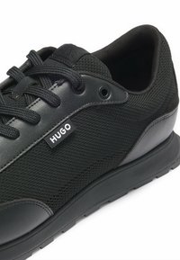 Scarpa sportiva nera con tomaia in rete e dettagli in pelle, dotata di lacci rotondi, logo laterale e suola in gomma texturizzata.
