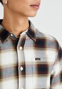Camisa de cuadros con tonos de rojo, blanco y gris. Diseño de botones con un bolsillo en el pecho y una pequeña etiqueta con el logo "Lee" en el bolsillo.