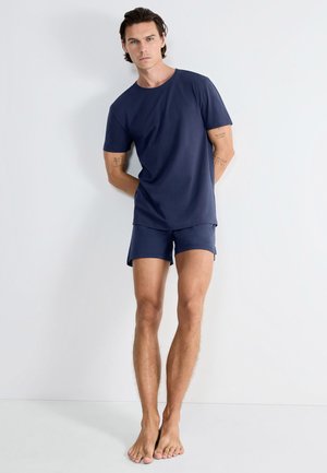 Man staat blootsvoets tegen een witte muur, draagt een marineblauw T-shirt met korte mouwen en bijpassende korte broek, armen achter de rug, neutrale uitdrukking.