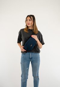 Borsa in tessuto blu navy a vita con chiusura a zip, forma rettangolare e un'etichetta con logo nera, indossata da una persona con una T-shirt grigia e jeans blu.