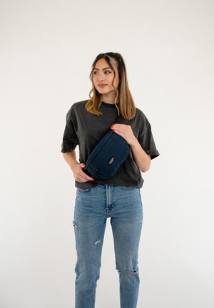 Marinblå tyg midjeväska med blixtlås, rektangulär form och en svart logotyp-lapp, bärs av en person i grå T-shirt och blå jeans.