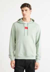 Joven vestido con una sudadera verde claro con un parche rojo del logo "HUGO" y pantalones negros, de pie contra un fondo blanco liso.