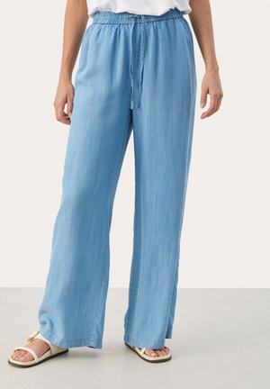 Lichtblauwe wijde broek met elastische tailleband en koordje, gedragen met witte sandalen tegen een effen achtergrond.