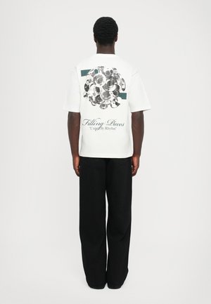 Witte t-shirt met een ronde grafische ontwerp van metalen platen op de achterkant, met de teksten "Filling Pieces" en "United By Rhythm". Zwarte broek.
