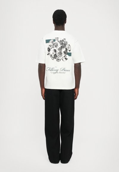 Witte t-shirt met een ronde grafische ontwerp van metalen platen op de achterkant, met de teksten "Filling Pieces" en "United By Rhythm". Zwarte broek.