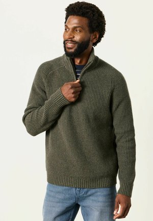 Mann trägt olivgrünen, gerippten Zip-up-Pullover und blaue Jeans, lächelt und schaut seitlich vor schlichtem Hintergrund.
