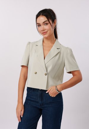 Νεαρή γυναίκα με σκούρα μαλλιά σε αλογοουρά, ντυμένη με ανοιχτό μπεζ κοντό σακάκι με κοντά μανίκια και σκούρο μπλε τζιν, χαμογελώντας.