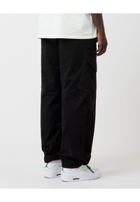 Persona que lleva pantalones cargo negros y zapatillas blancas con acentos verdes, de pie frente a un fondo claro y liso.
