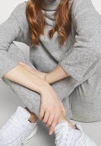 Szary, teksturowany sweter i pasujące spodnie, w połączeniu z białymi sneakersami. Ubranie ma wysoki kołnierz i luźny krój.