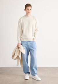 Beige gerippter Pullover, hellblaue gerade geschnittene Jeans und weiße Sneaker. Hält eine beige Jacke. Einfaches, lässiges Outfit vor einem neutralen Hintergrund.