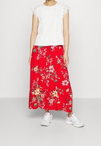 Top blanco de manga corta con detalles de encaje, combinado con una falda larga roja con flores que presenta acentos en amarillo y verde, y zapatillas deportivas blancas.
