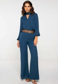 Mono azul verdoso con mangas largas, escote en forma de llave y pantalones de pierna ancha. Un cinturón marrón detallado en la cintura añade textura y contraste.
