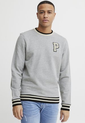 Grauer Sweatshirt mit einem gerippten schwarzen und cremefarbenen Kragen und Bündchen. Verfügt über ein großes gesticktes "P" auf der Brust. Trägt man über hellblauen Jeans.