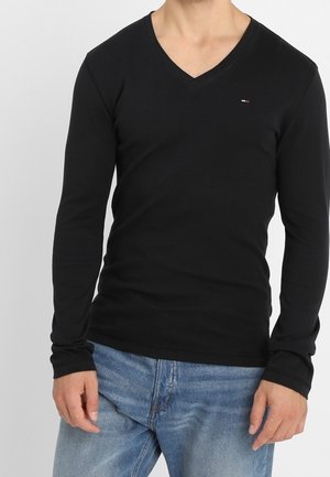 Zwarte longsleeve V-hals shirt van zacht materiaal, met een getailleerd ontwerp en een klein logo-accent op de borst. Gecombineerd met blauwe jeans.