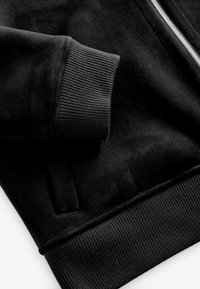 Jachetă din fleece negru cu manșete și talie ribbate. Are închidere cu fermoar și material moale, texturat. Detaliu cu buzunare laterale vizibile.
