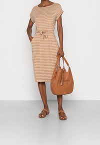 Frau mit einem beigen und weißen gestreiften knielangen Kleid mit gebundenem Taillenbereich, die eine braune Lederhandtasche hält und braune Sandalen trägt.