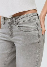 Sivi denim hlače s slim fit krojem, ki vključujejo srebrne okrasne žebljičke ob strani, klasične žepke in gumbasto zapiranje v pasu.