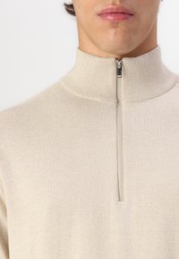 Suéter beige con cremallera y cuello alto, que presenta un diseño ajustado y ribetes acanalados, hecho de una tela suave y texturizada.