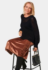 Zwarte gebreide trui, metallic bronzen geplooide rok, zwarte hoge laarzen, een zwarte handtas met gouden accenten vasthoudend, zittend op een kruk.